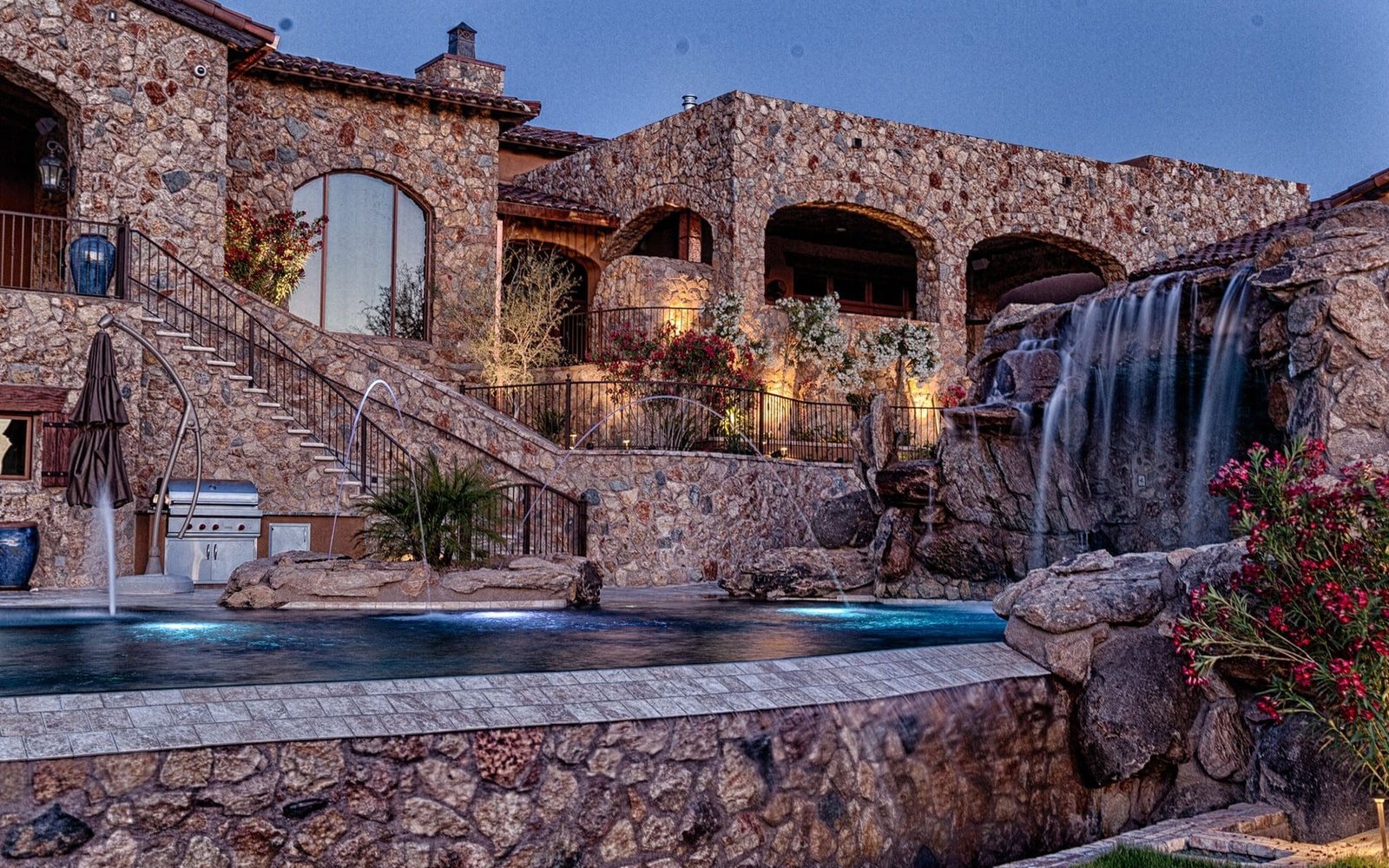 Stone estate pool with lit waterfall and raised spa at dusk, built by No Limit